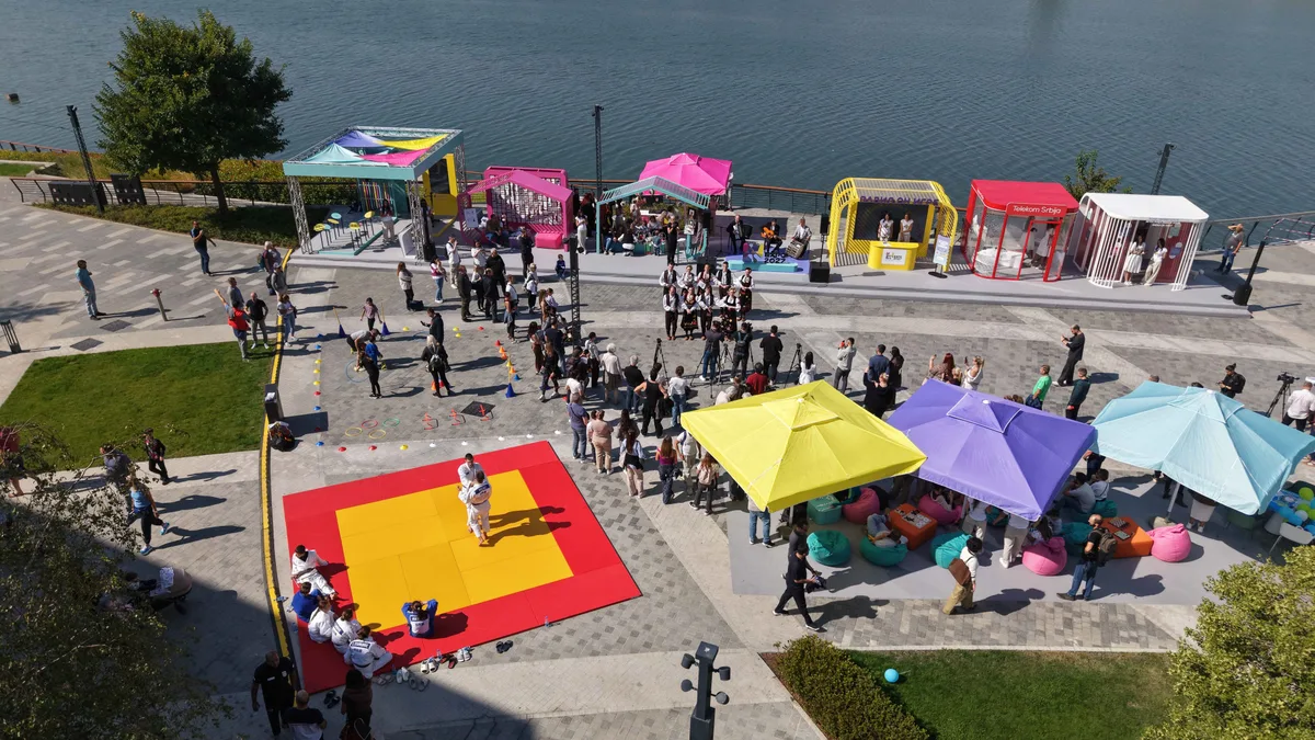 Drone view of Sava Promenada in Belgrade: a crowd around a red and yellow mat where judoka in white give a demonstration, a folk ensemble in traditional costume by a small stage, colourful stands and parasols, and the Sava river behind